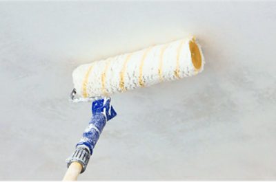 A close-up view of white paint being applied to a ceiling using an extended paint roller.