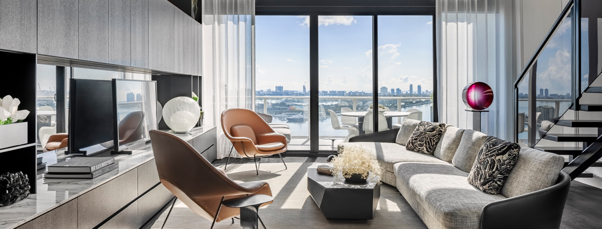 Modern urban loft living area with postmodern sectional and twin leather armchairs in front of flat screen TV with a view to an outside balcony area overlooking a city skyline.