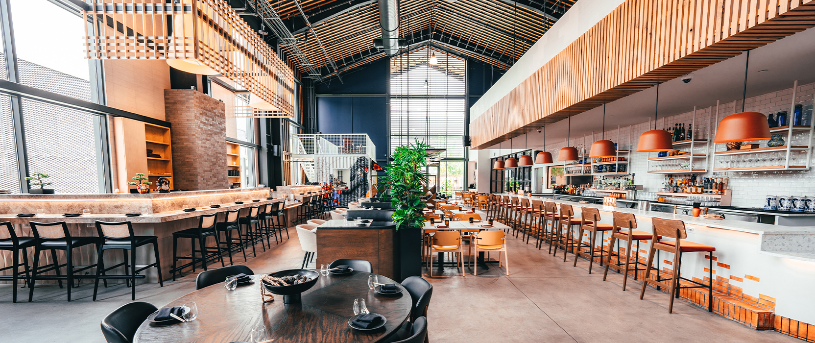Interior of spacious and high-ceilinged food hall with bar seating and tables throughout, natural wood accents, brick, and dark blue and white color scheme.