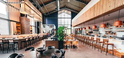 Interior of spacious and high-ceilinged food hall with bar seating and tables throughout, natural wood accents, brick, and dark blue and white color scheme.