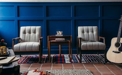 Two gray tufted chairs with wood fraims and a shared accent table situated on woven rugs in front of a board-and-batten wall in a rich blue color, with an acoustic guitar on a stand off to the side.