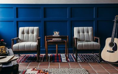 Two gray tufted chairs with wood frames and a shared accent table situated on woven rugs in front of a board-and-batten wall in a rich blue color, with an acoustic guitar on a stand off to the side.