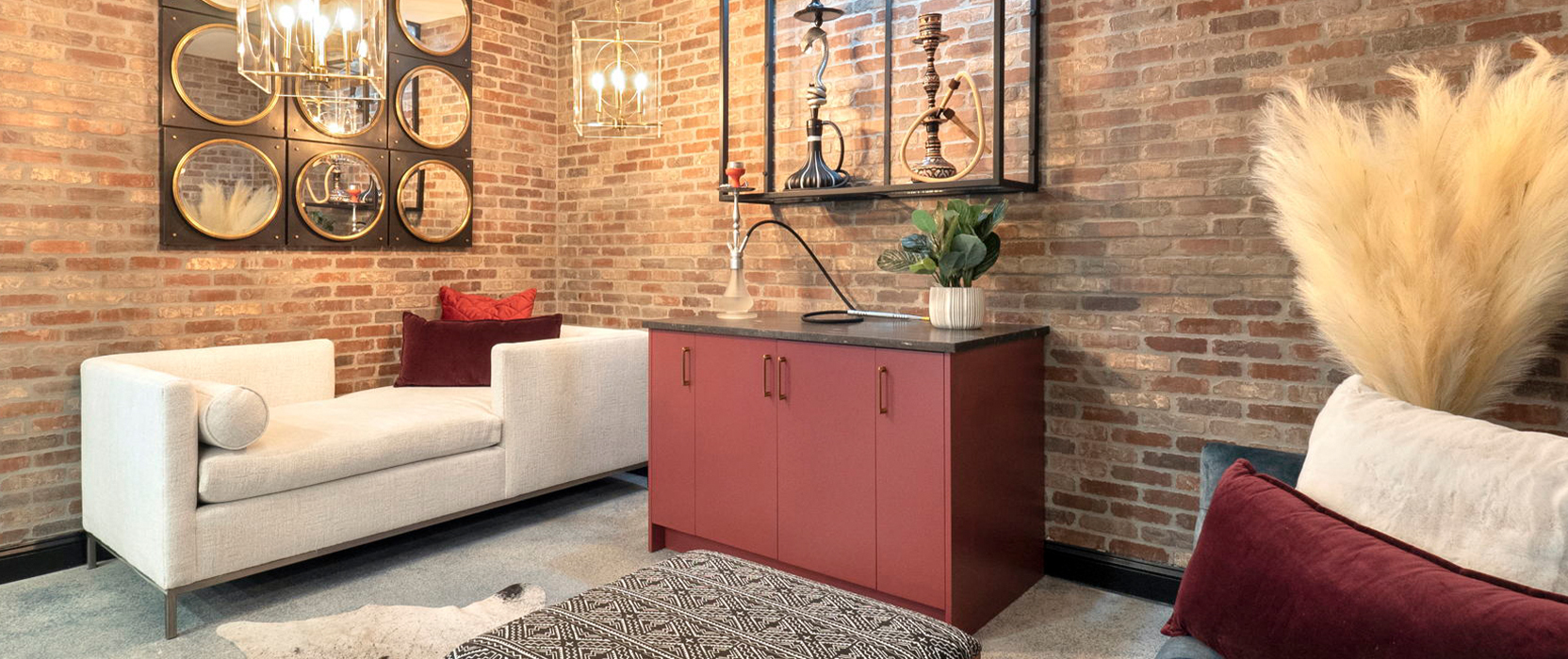 Hookah bar area in basement with mirrored wall art on brick walls, a red painted bar, hookah pipes on display and chaise lounge style seating.