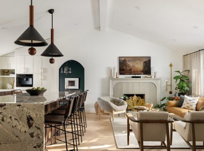 A modern open-concept living space with a kitchen bar, pendant lighting, vaulted ceilings, and a cozy living room with a fireplace and landscape painting.