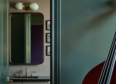 A glimpse through a bathroom door showing a vanity with a mirror, sconce, and hanging art, with a partial view of a string instrument in the foreground. 