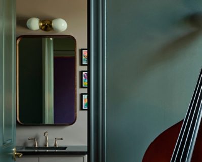 A glimpse through a bathroom door showing a vanity with a mirror, sconce, and hanging art, with a partial view of a string instrument in the foreground. 