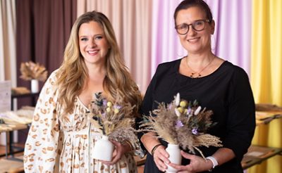 Two women smile holding their dried-flower bouquets in small white vessels in front of a curtained backdrop in various Color Capsule of the Year hues.