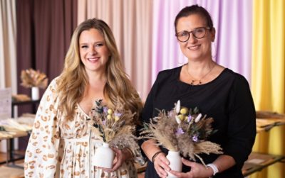 Two women smile holding their dried-flower bouquets in small white vessels in front of a curtained backdrop in various Color Capsule of the Year hues.