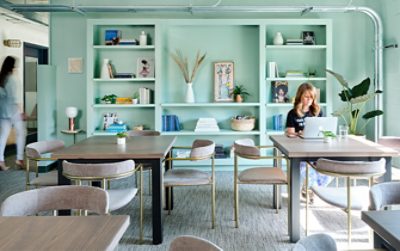 Interior of the Lola coworking space, with light blue-green walls and built-in shelving full of books and decor, with a woman working on a laptop at one of two modern worktables in foreground.