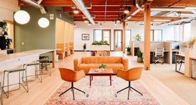 Open concept office space with light wood flooring, exposed wood beams, kitchen counter under globe lights and walled-off booths at left, sofa and chairs in center, and cubicle workspaces to the right.