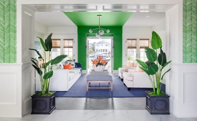 Living room with white sofa and armchairs, blue rug, and white walls and ceiling with bright green painted stripe down the center of the room, twin potted palms in foreground.