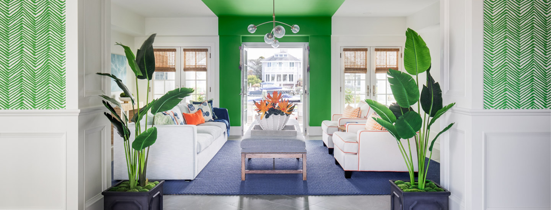 Living room with white sofa and armchairs, blue rug, and white walls and ceiling with bright green painted stripe down the center of the room, twin potted palms in foreground.