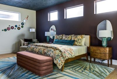 An eclectic bedroom with floral bedding, three high windows, a botanical patterned rug, and an accent wall painted Sherwin-Williams Rojo Marrón.