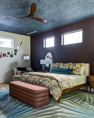 An eclectic bedroom with floral bedding, three high windows, a botanical patterned rug, and an accent wall painted Sherwin-Williams Rojo Marrón.