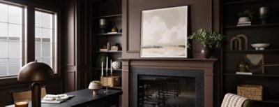 A dark study color-drenched in Sherwin-Williams Chateau Brown with a desk, built-in shelves, fireplace, large landscape painting, and houseplants.