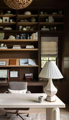 A split view of a warm, traditional home office in the chocolate box style with dark built-in shelves and walls painted Sherwin-Williams Sable, a light desk, gray office chair, and sculptural table lamp.