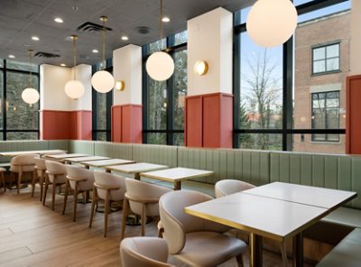 Modern restaurant interior with white tables, light green banquettes around perimeter, beige chairs, large windows, and round pendant lights. 
