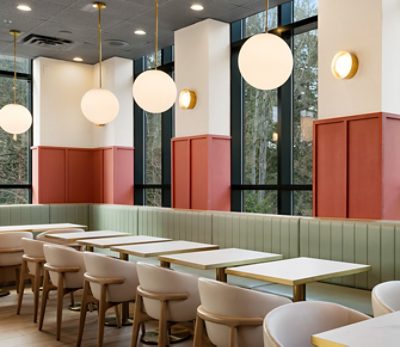 Modern restaurant interior with white tables, light green banquettes around perimeter, beige chairs, large windows, and round pendant lights. 