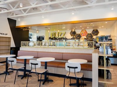 Interior of Better Buzz café with modern decor, featuring tables, a cushioned bench, and hexagonal panels. "BUZZ" is prominently displayed on the back wall. 