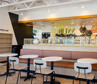 Interior of Better Buzz café with modern decor, featuring tables, a cushioned bench, and hexagonal panels. "BUZZ" is prominently displayed on the back wall. 