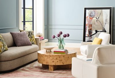 A Dutch Tile Blue living room with neutral furnishings, a burl wood coffee table, and a large abstract art piece leaning against the rear wall.