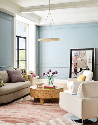 A Dutch Tile Blue living room with neutral furnishings, a burl wood coffee table, and a large abstract art piece leaning against the rear wall.
