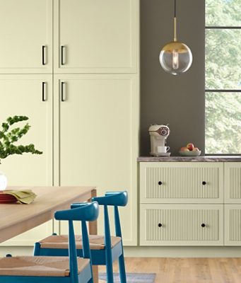 A modern kitchen with Celery painted cabinets, a wooden table with Georgian Bay blue chairs, and an espresso maker on marble countertops against a wall painted Sherwin-Williams Cocoon.