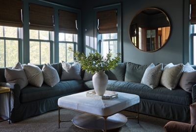 A cozy living room in tones of dark gray, including an inviting sectional sofa full of neutral-colored pillows, a coffee table with a vase of greenery, Homburg Gray walls, and natural light from the surrounding windows.