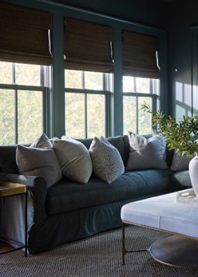 A cozy living room in tones of dark gray, including an inviting sectional sofa full of neutral-colored pillows, a coffee table with a vase of greenery, Homburg Gray walls, and natural light from the surrounding windows.