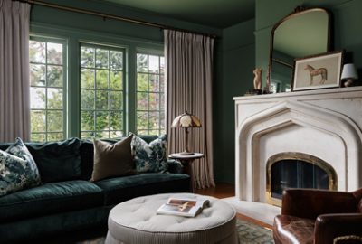 Cozy living room with a dark velvet sofa, round ottoman, and marble fireplace, illuminated by large windows with beige curtains and drenched in a dark green paint color.
