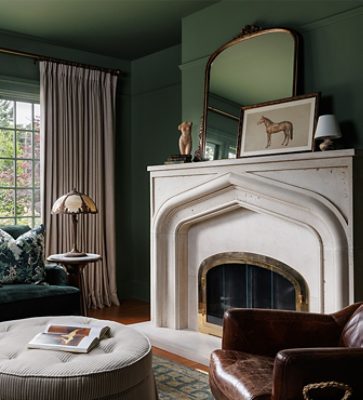 Cozy living room with a dark velvet sofa, round ottoman, and marble fireplace, illuminated by large windows with beige curtains and drenched in a dark green paint color.