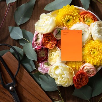 A white bowl full of flowers with eucalyptus leaves on the dark wood table holding the bowl. A color chip for Navel is on top of the flowers.