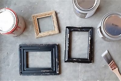 Three picture frames, three closed paint cans, and a paintbrush arranged on a concrete surface, suggesting a DIY painting project