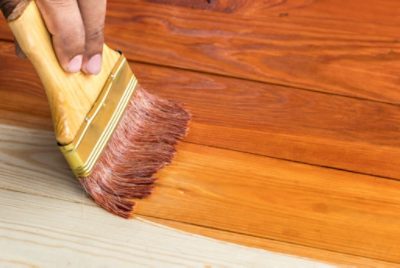 Close-up of a person using a brush to apply stain to a wood floor.
