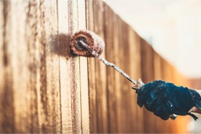 Close-up of a person wearing gloves using a roller to apply wood stain to a wooden fence.