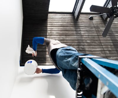 Una persona parada en una escalera sosteniendo un balde de pintura blanca mientras pinta una pared.