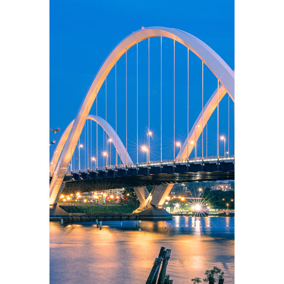 An illuminated bridge at night, reflecting on the water, leading to a brightly lit city.