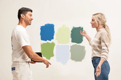 Two people looking at six different colored paints painted out on a tan wall.