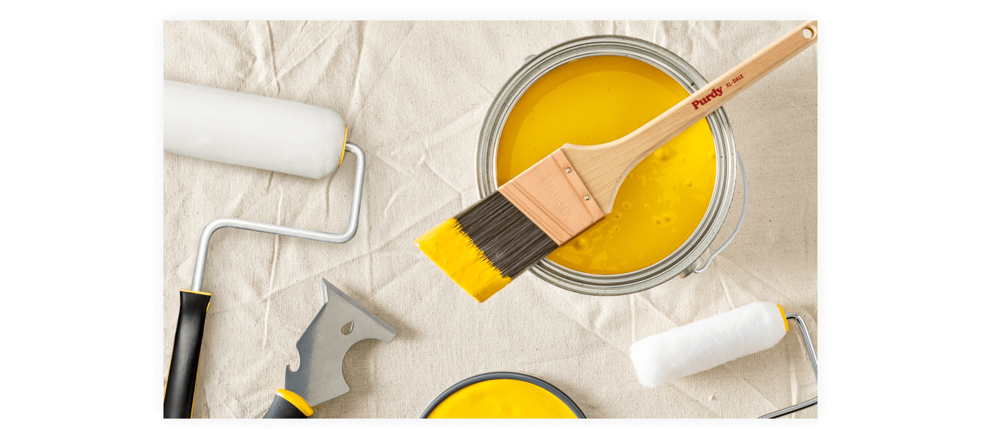 A Purdy paint brush set across an open can of yellow paint with other supplies nearby on a drop cloth.