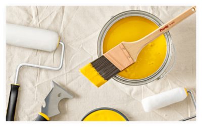 A Purdy paint brush set across an open can of yellow paint with other supplies nearby on a drop cloth.