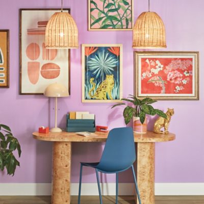 A desk and chair in front of a wall painted a light purple with many framed prints hung.