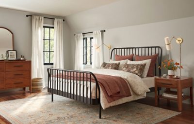 A large bedroom with lots of natural light with a large bed with black rails, wooden nightstand and wooden dresser with a mirror.