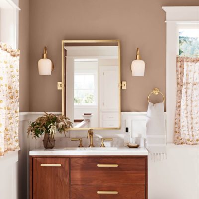 A brightly lit bathroom with a sink with wooden cabinets and white counter top and a mirror with gold trim against a tan wall. 