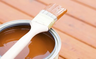 An open gallon of brown deck paint and a paint brush.