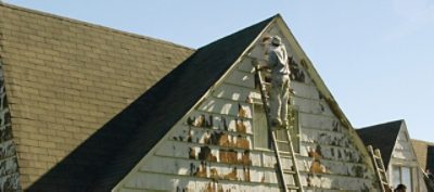 A paint professional is standing on a ladder and sanding off paint from an exterior home.