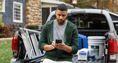 A person sitting in the back of a truck on his phone with paint supplies in the background.