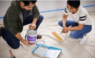 Two people painting, one pouring and another with a paint roller over a tarp.