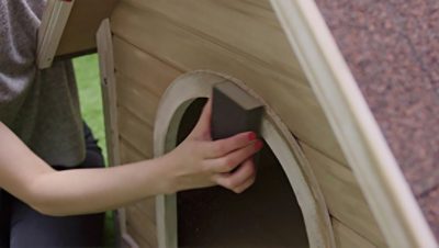 A woman sanding a dog house with a sanding block. 