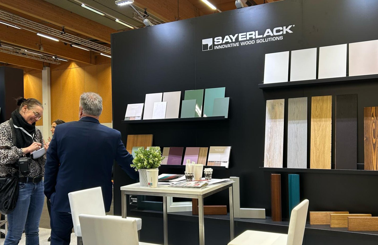 Sayerlack booth at a trade show with wood finish samples, brochures, and two people interacting near a display wall.