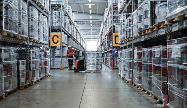 Sayerlack warehouse with stacked metal coating containers and worker using pallet jack in central aisle.